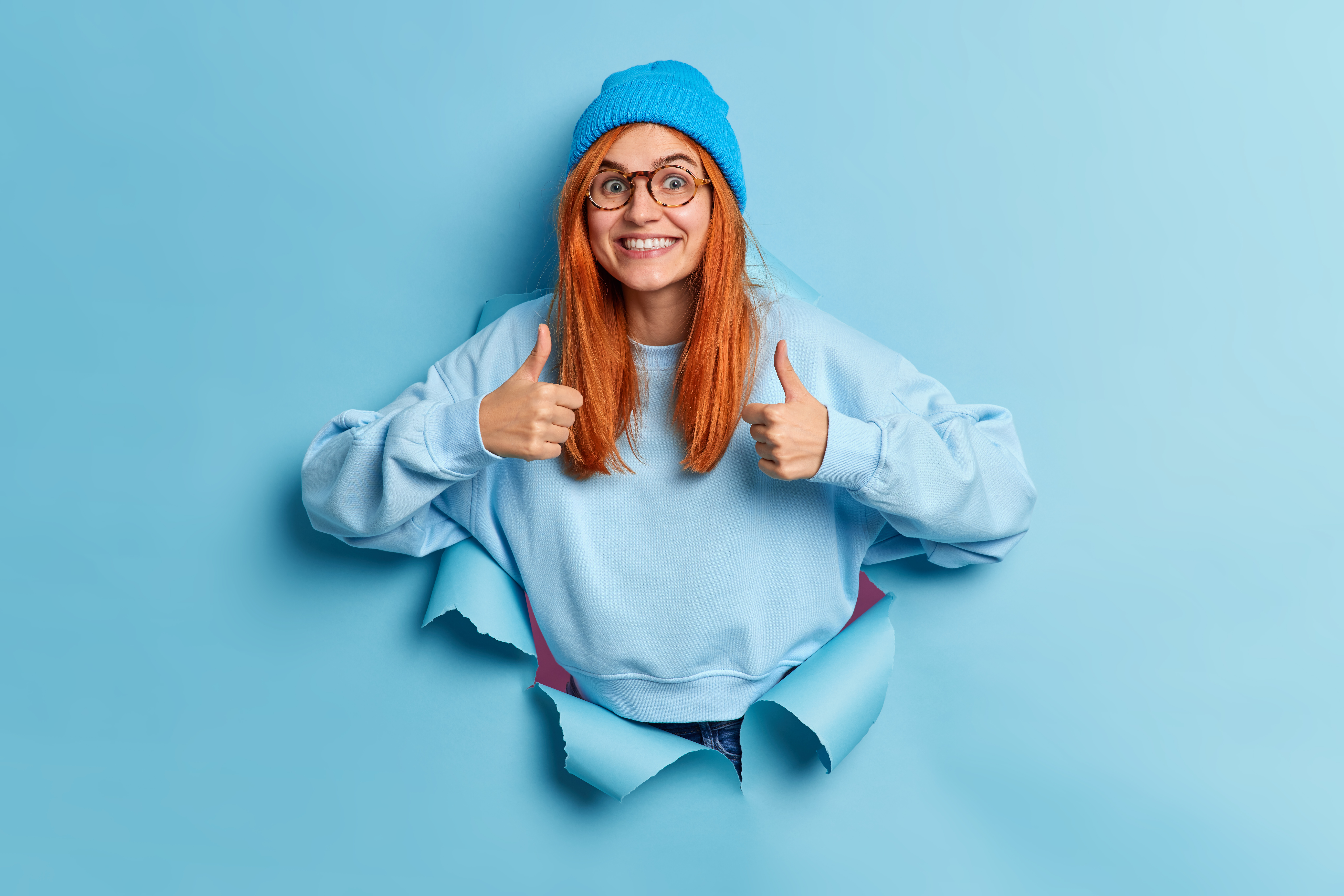 cheerful-redhead-european-woman-makes-thumb-up-gesture-makes-excellent-sign-approves-something-smiles-broadly-wears-hat-sweater-breaks-through-paper-hole