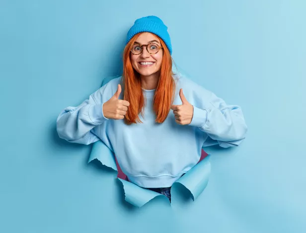 cheerful-redhead-european-woman-makes-thumb-up-gesture-makes-excellent-sign-approves-something-smiles-broadly-wears-hat-sweater-breaks-through-paper-hole